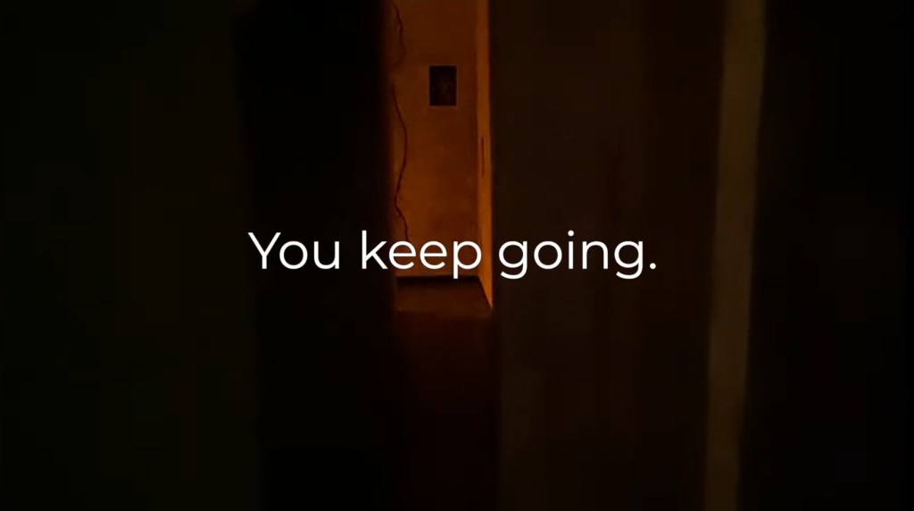 A dark picture down a narrow hallway, lit only by an amber light coming from around the corner at the other end. A vine and a poster can also be seen. White words in the center read "You keep going."