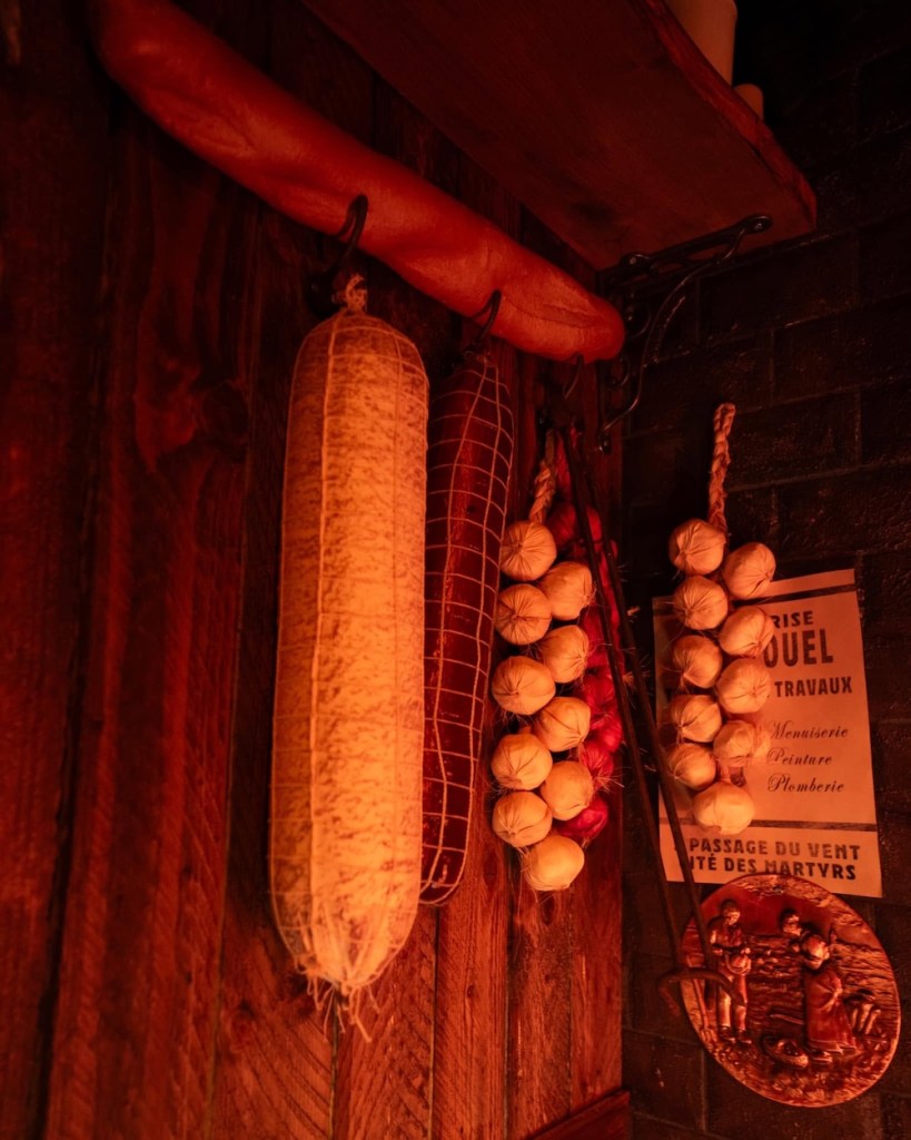 a baguette, cloves or garlic, and cured meat hanging in a shop against a wood paneled background, under a shelf