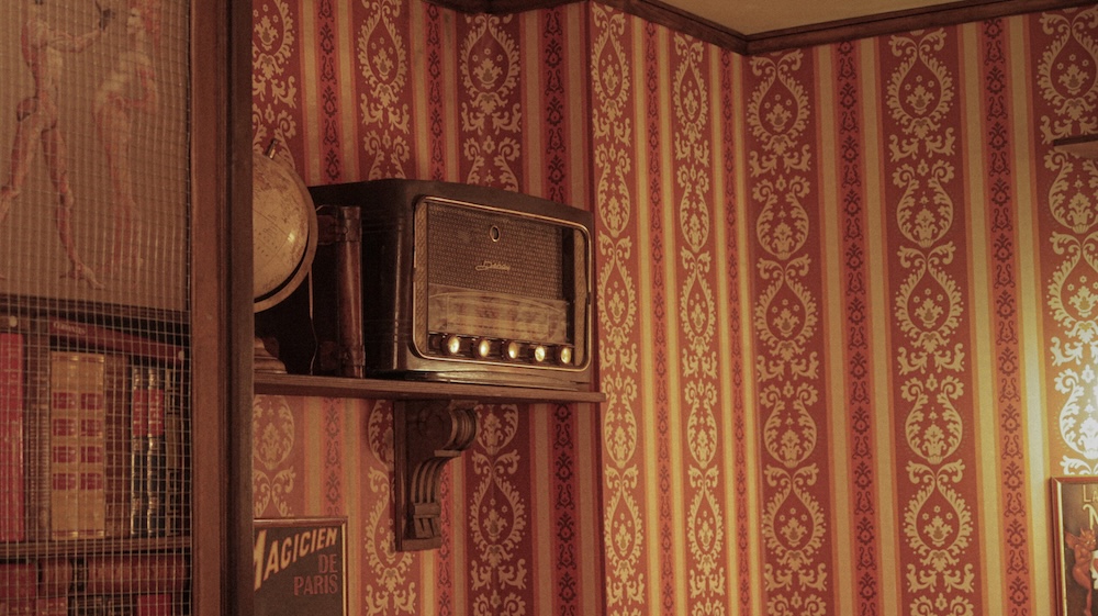 an old timey radio sets on a wooden wall shelf against walls covered in red patterned and striped wall paper, and on the left side of the radio is a bookshelf
