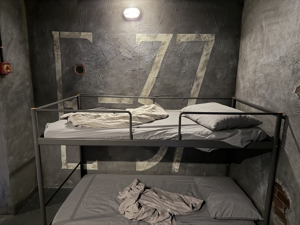 A concrete and brick walled jail cell with "E-77" pained behind bunk beds.