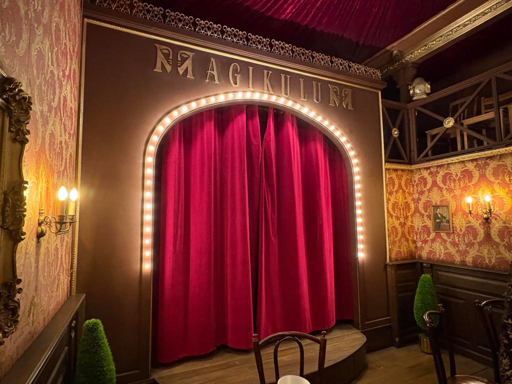 Image of the Magikulum theater, with a red velvet curtain, white lights around the stage opening, red & gold wallpaper, and a few chairs for the audience