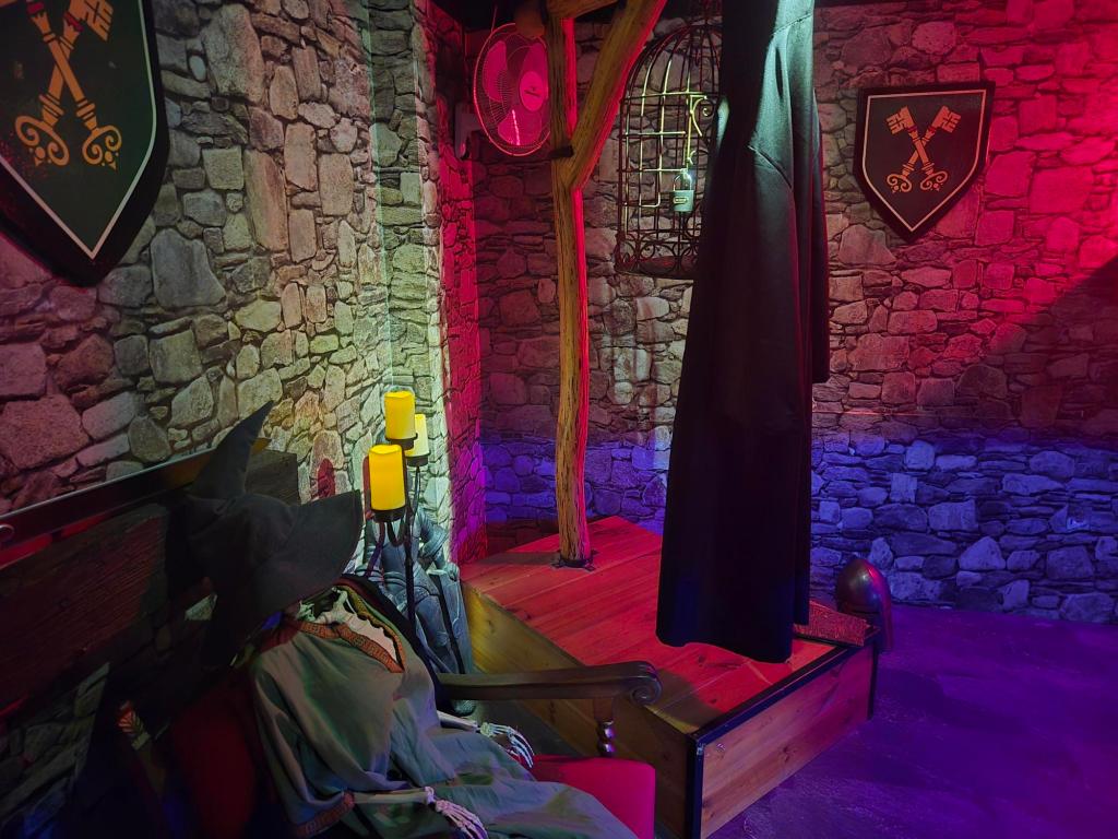 Stone room with a gallows that has a metal bird cage and a black robe hanging from it. A shield with crossed keys painted on it hangs from the wall. A skeleton with a wizards hat and robes is sat in a chair on the left side.