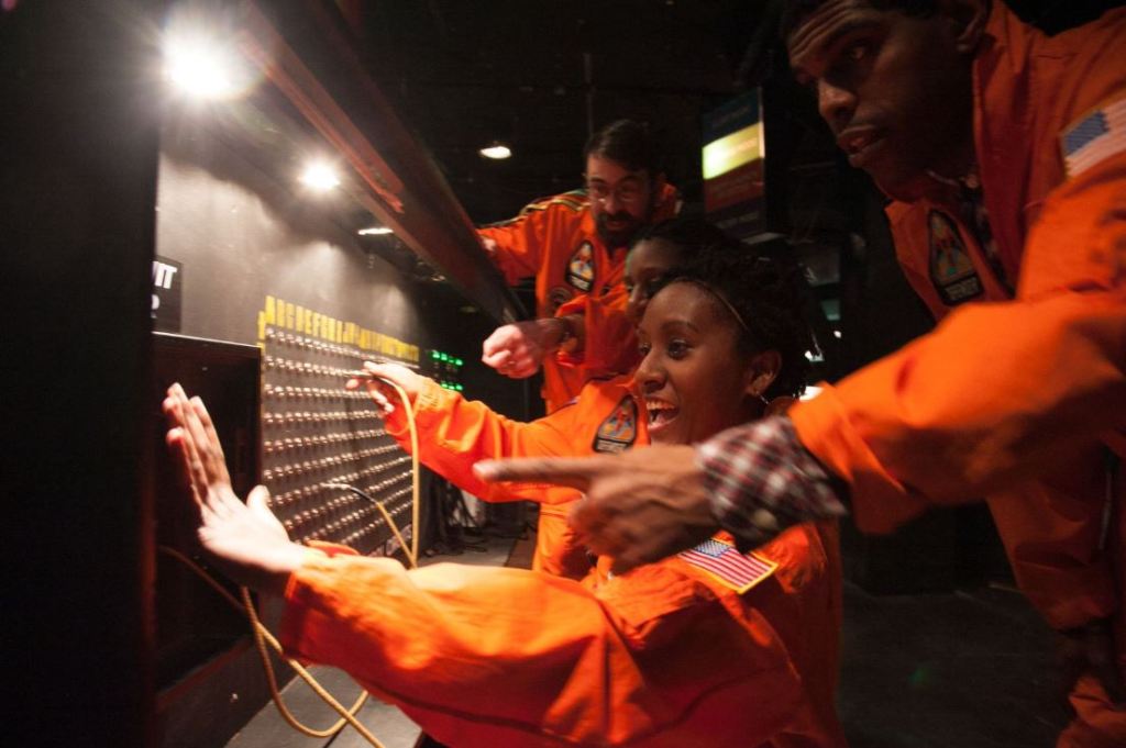 Group of people in orange jumpsuits gathered around a wall with many long rows of switchboard jacks.