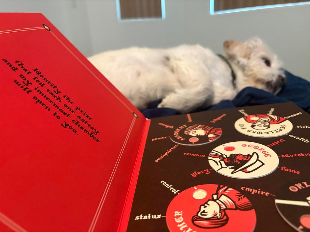 A picture of an opened book. The left page is red and features the text, "Identify the prize that led each one astray and my innermost chamber will open to you." The right of the book features 5 cardboard dials, each with a red, black, and white linocut illustration of a person and their name in an ornate font. The dials each can rotate to point to five words, such as "prestige," "judgment," and "deathlessness." In the background, there is a white terrier lying in bed.