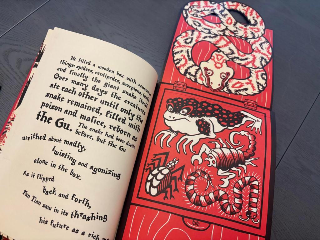 An open book on a grey table. The left side of the book features stark black renaissance-style text describing a snake writing in a box; the text is typeset to mimic the snake's movements, and several lines are curved in subtle arcs. The right side of the book contains a linocut illustration of a red wooden box filled with white, red, and black creatures, including a scorpion, frog, beetle, and centipede. The box is made of sturdy cardstock and folds out, the crease line depicting the hinges of the box. Sitting on the lid of the box is a curled snake with black and red markings.