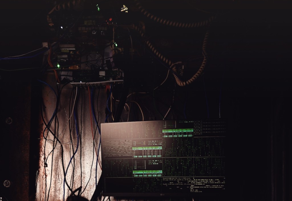 A wall covered in wires and technology with a screen displaying a large volume of green text.