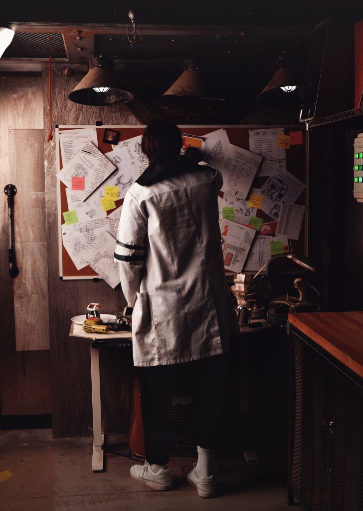 A person in a lab coat in a dirty lab, with a bulletin board covered in papers.