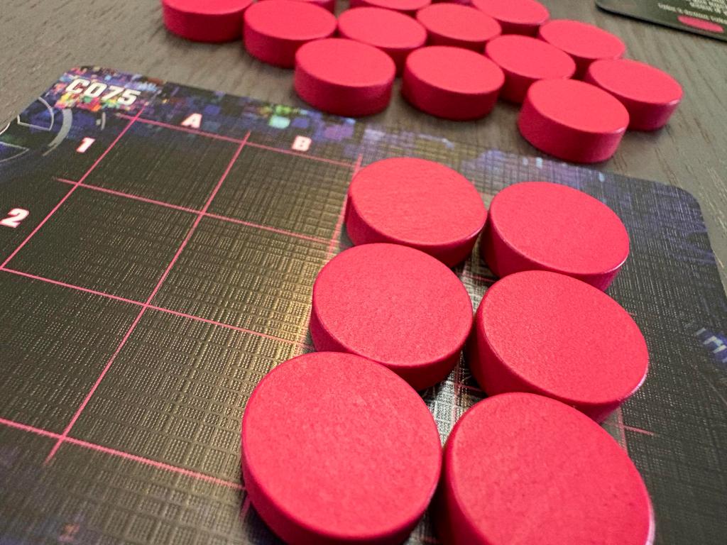 Six red, wooden tokens sit on a card. Behind them are over a dozen additional tokens sitting on a grey table.