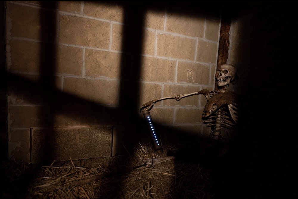 A skeleton sitting against a wall in a jail cell. It's hand holds a glowing tube.
