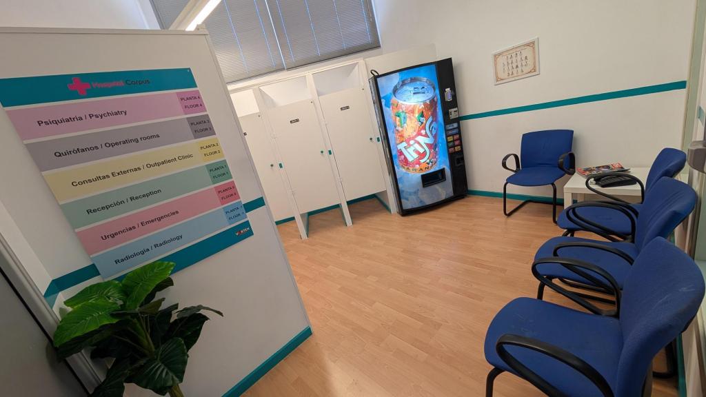 A clinical hospital waiting room, with a list of floors, blue chairs, and a vending machine