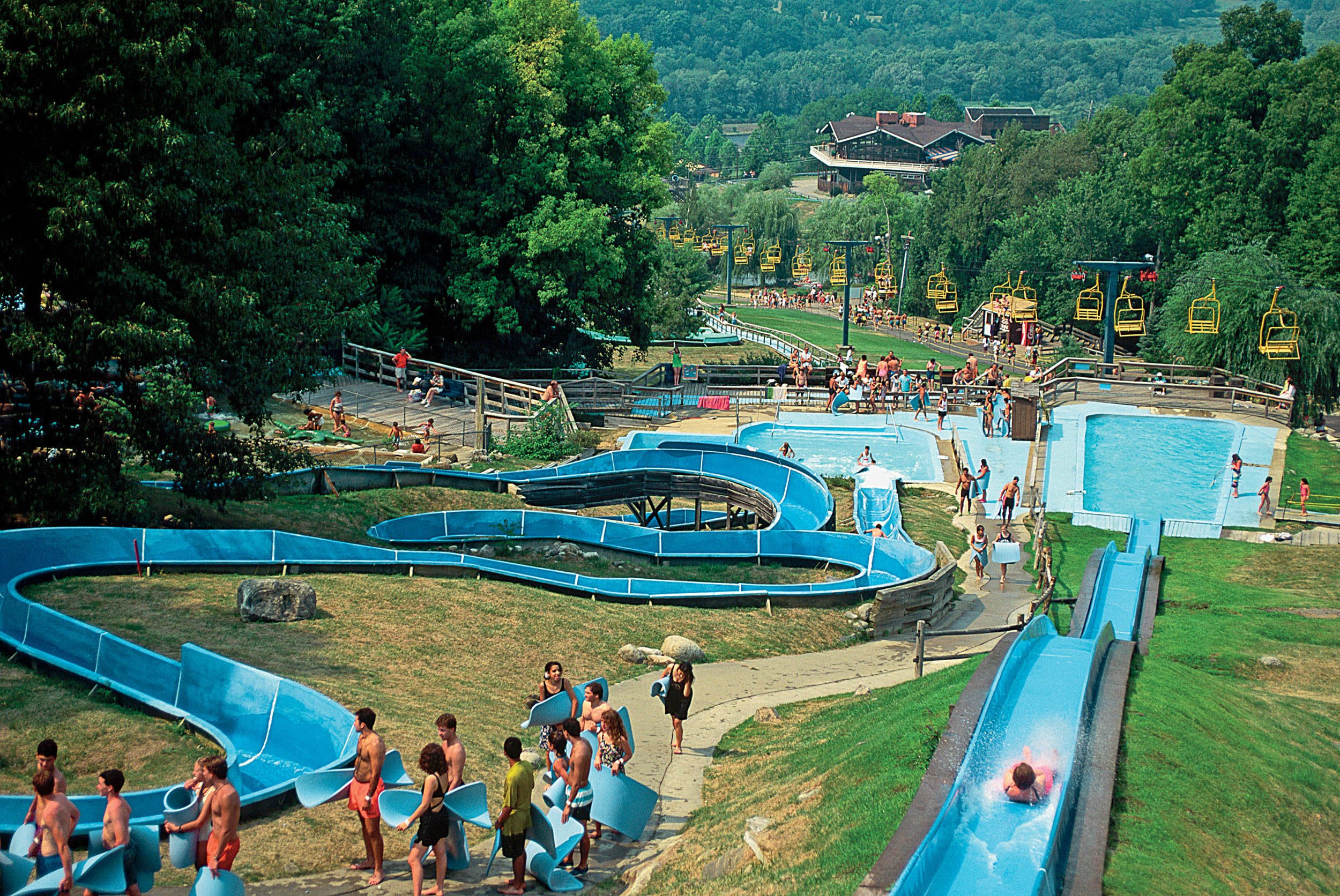 people holding mats lined up for water slides. background of pools and ski lifts