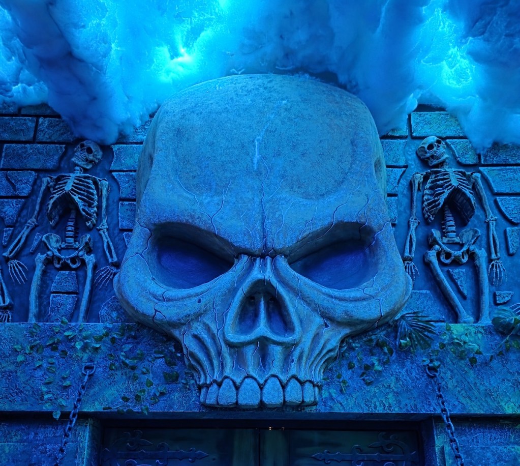 The exterior wall of a castle with a massive stone skull on its face. It is flanked by skeletons. Above is cloud cover.