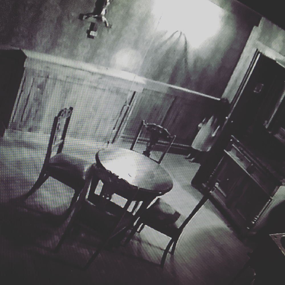 View of a kitchen in an old house through a grainy black and white camera. A young girl with long dark hair covering her face stands in a doorway.