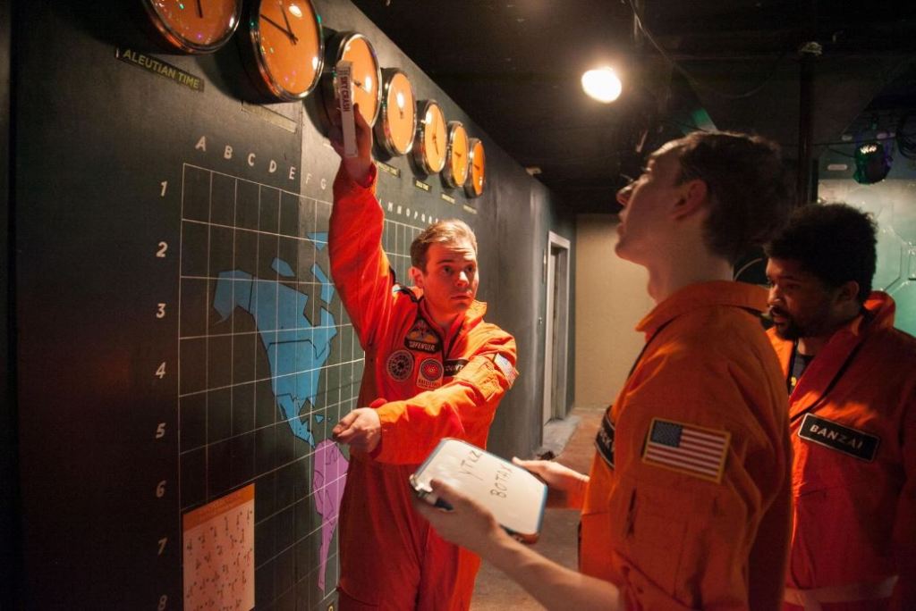 3 men in orange coveralls looking at map and clocks on a wall. One of the men is holding his arms in the positions of the clock hands.