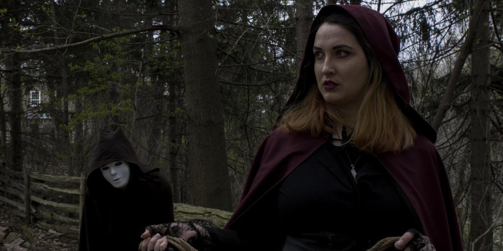 A masked figure in the woods following a woman in a maroon cloak