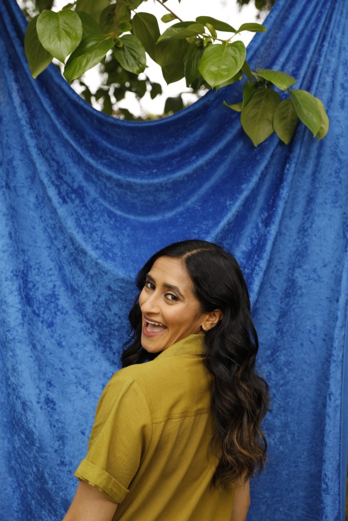 southeast asian woman with long wavy dark hair wearing a mustard color blouse, looking back over her shoulder and smiling
