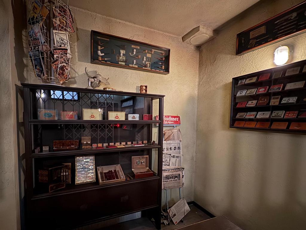 Wide angle showing several display cases containing cigars and other tobacco products, a newspaper stand next to it, and a magazine rack on top.