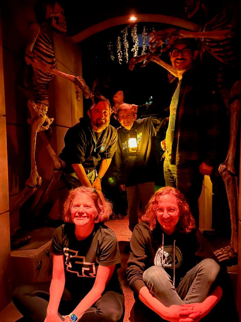 Team photo inside a Hidden Temple set with skeletons on the walls with 2 women sitting on the ground in the front, 2 men and a boy standing behind them, and a teenager barely visible in the back.