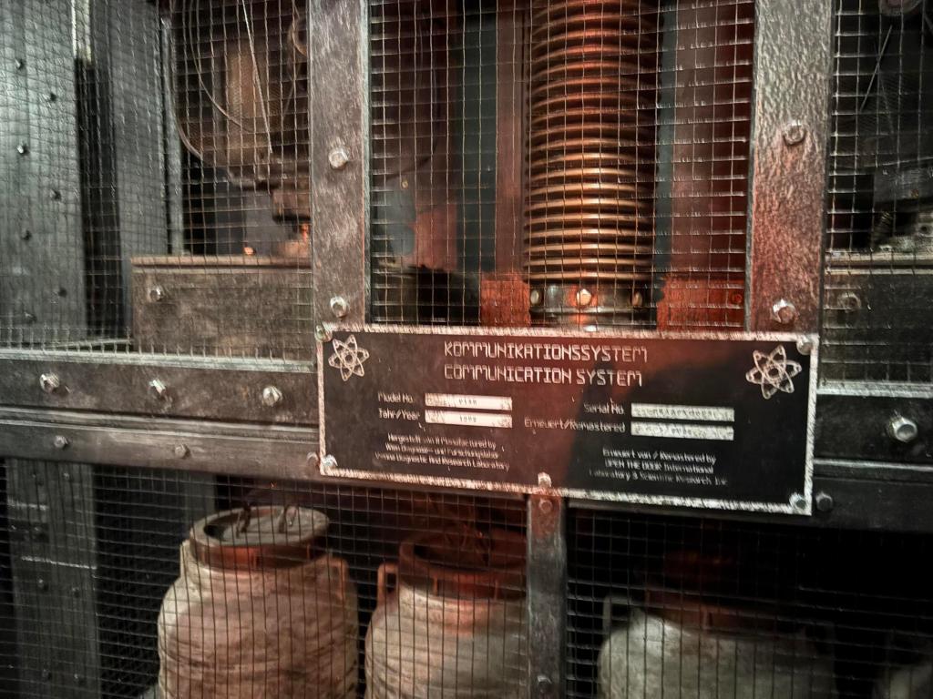 Rustic wire mesh storage unit labeled Communication system in English and German. Several canisters and mechanisms are on display.