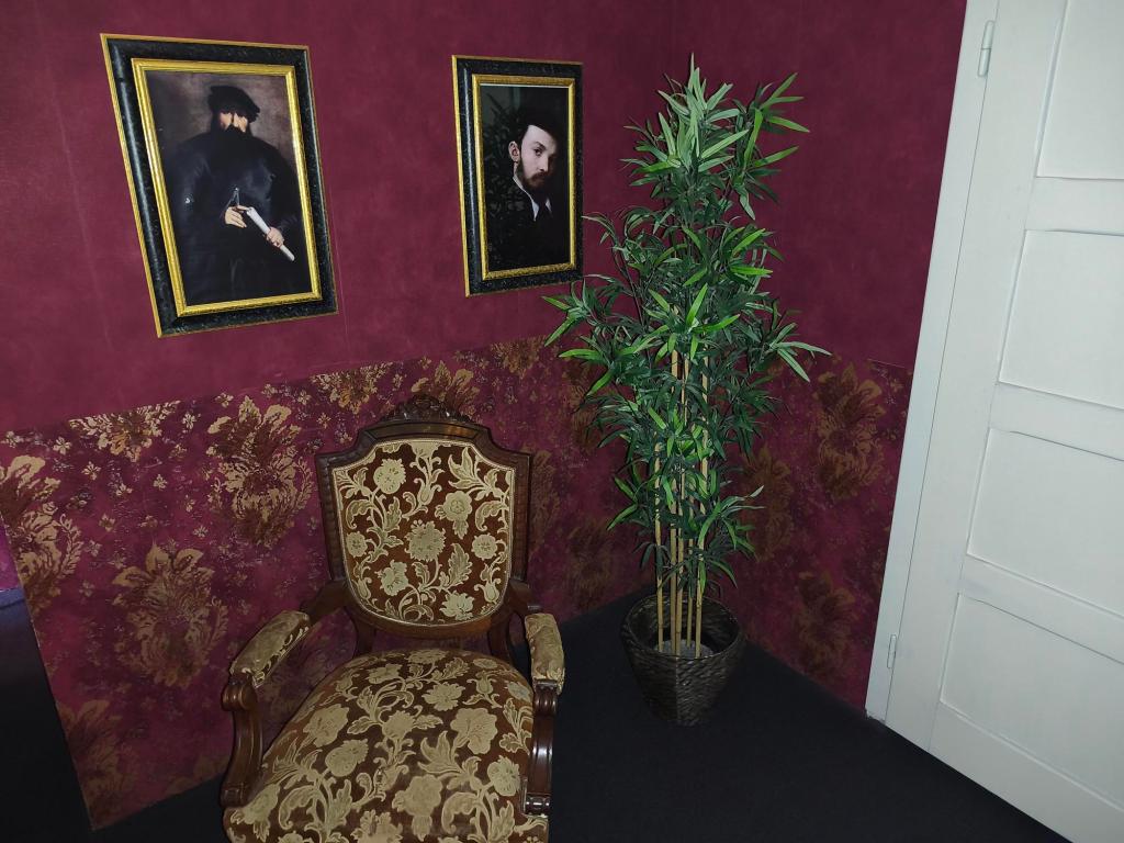 Room with dark maroon and gold wallpaper and a maroon and gold upholstered chair. Two portraits of serious-looking men hang on the wall.