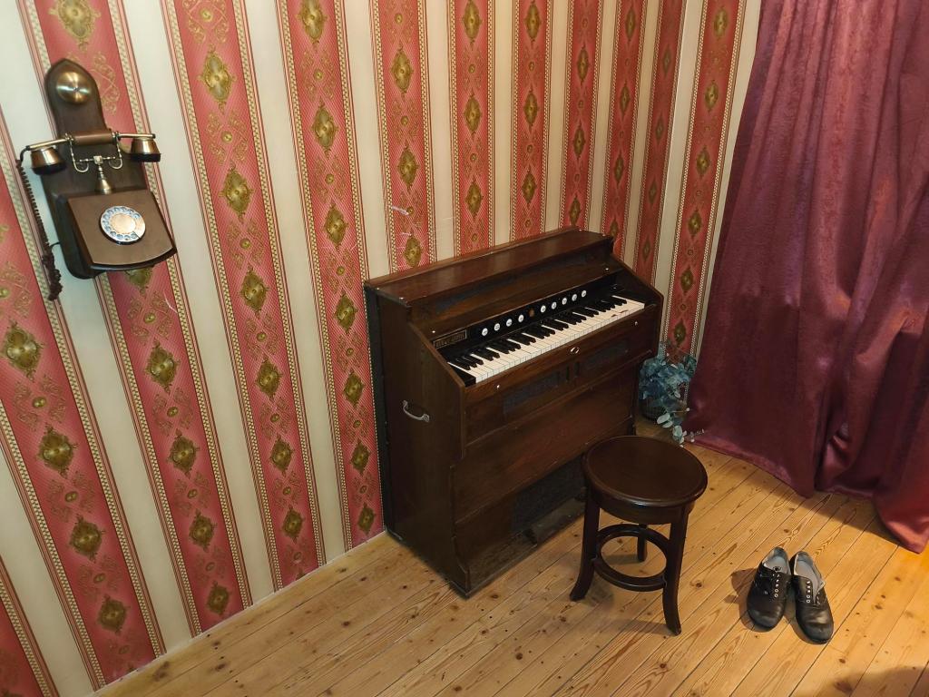 Image of a room with pink and white striped wallpaper, a small piano, and an old-fashioned wall phone. A pair of floppy shoes sit on the floor nearby.