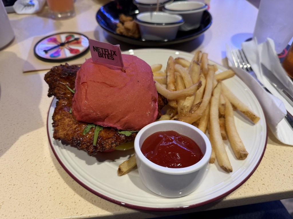 A chicken sandwich with a pink bun, fries, and ketchup. The sandwich has a flag that reads, "Netflix Bites" stuck in the bun.
