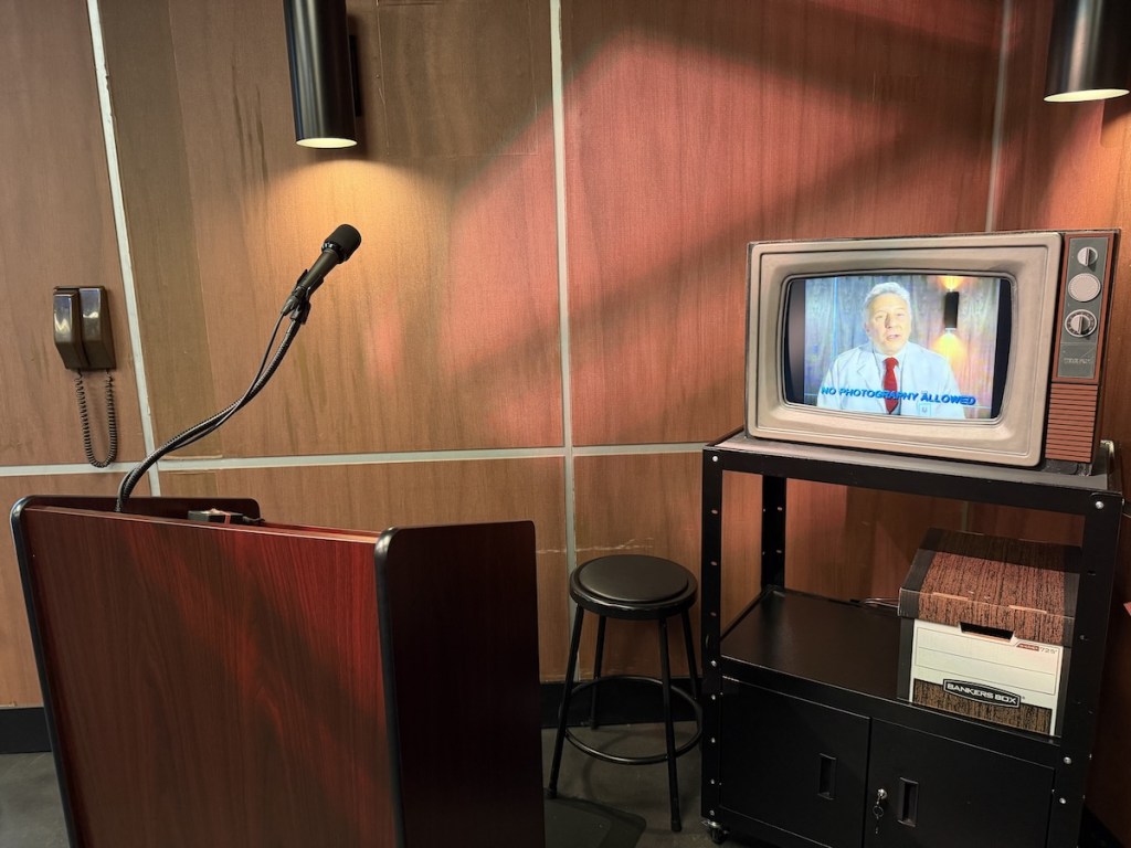 A 1980s, wood-paneled room with a podium, and an old TV that has a man on it. Text on the screen reads, "No photography allowed."