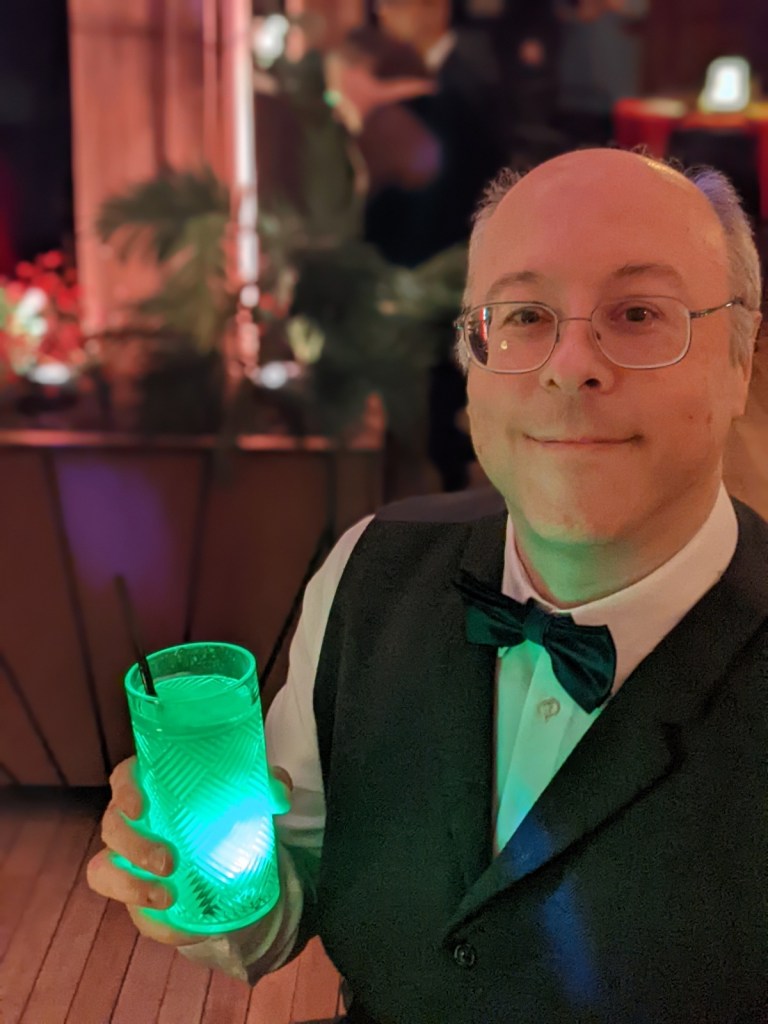 man with silver hair and glasses in a tuxedo vest and bowtie, holding a glowing drink