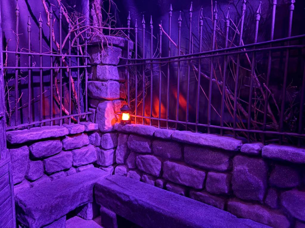 Image showing a corner wall made of rock, with a metal fence for the top portion. A single lantern lights the corner. Image has a violet overtone.