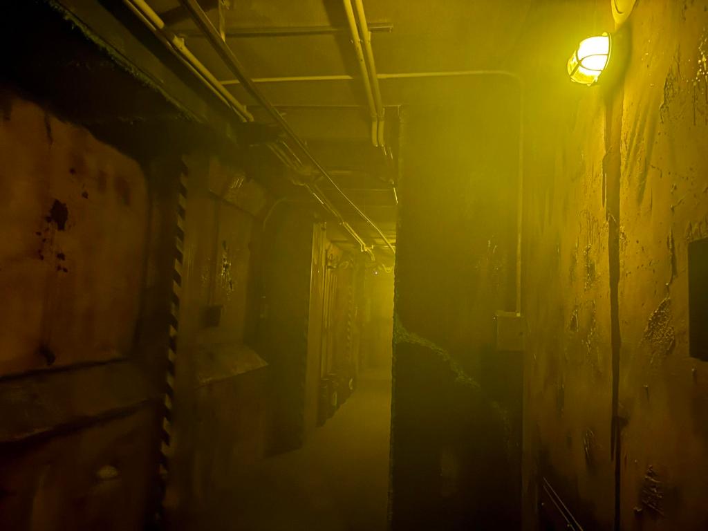 A long, smoke-filled hallway, with walls of stucco, exposed pipes, and just enough lighting to cast a yellow glow to the space, .