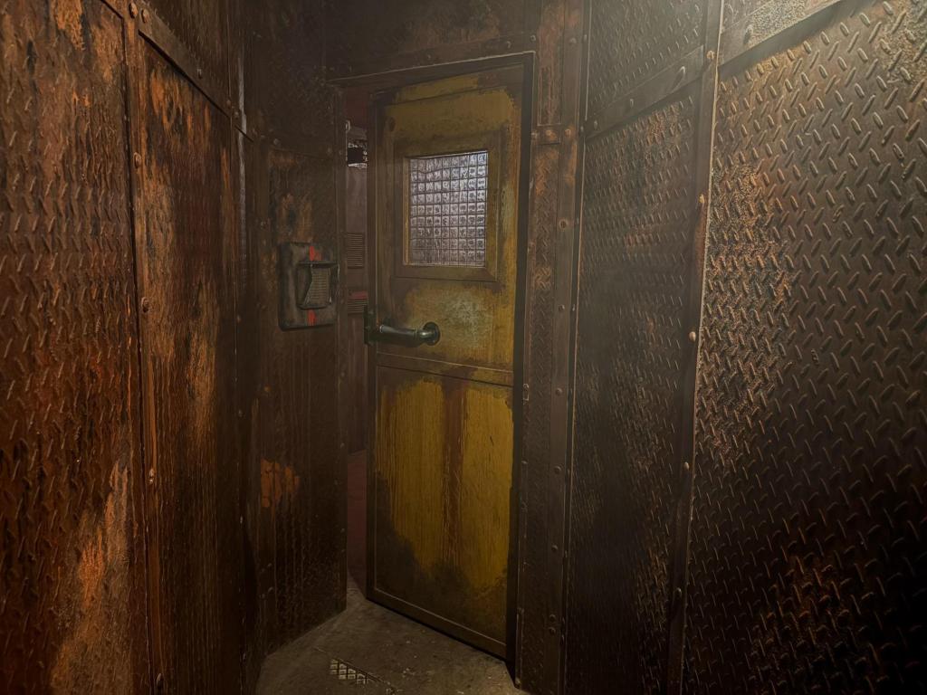 Image of rusted, stained metal walls leading to a stained yellow door. The door is ajar, and there's an intercom next to it.