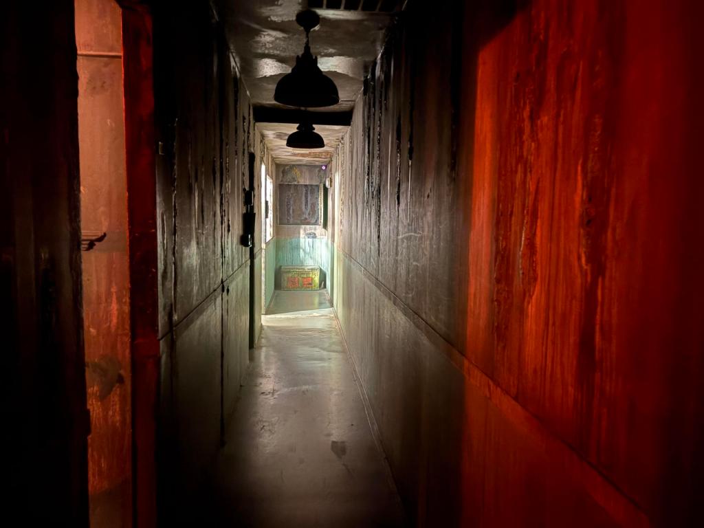 Long, darkened hallway with a room on the left, casting light into the narrow passage.