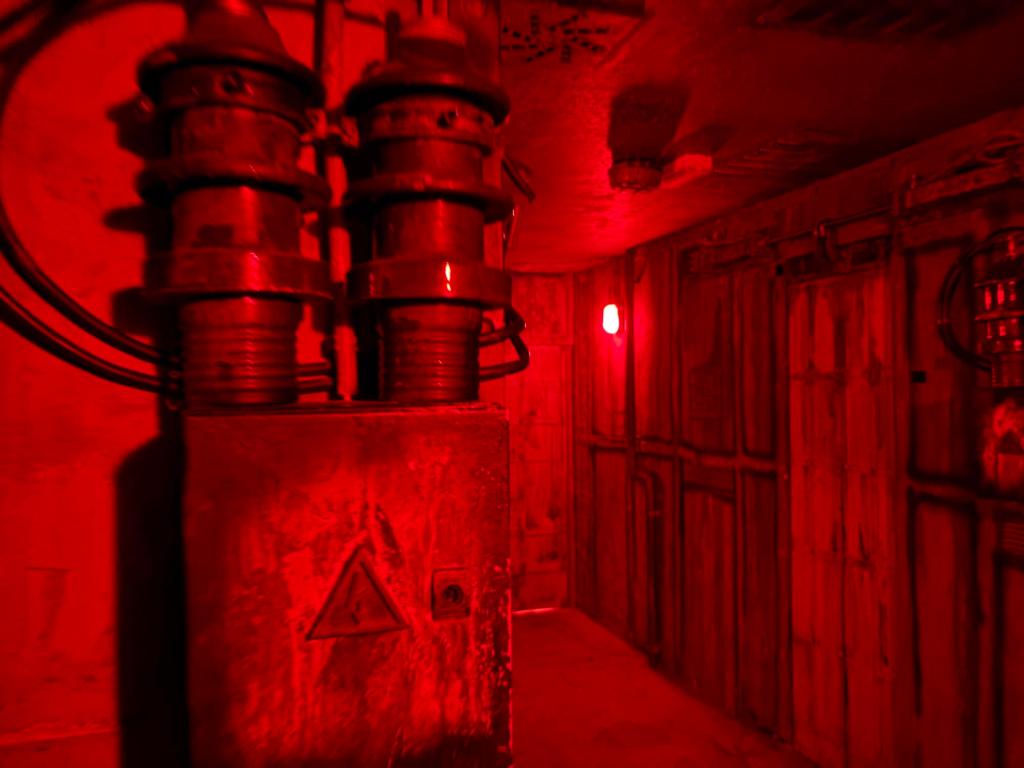 Closeup of a power box and cables, beyond which you can see a hallway lit by an industrial wall lamp. The scene is basked in deep red light.