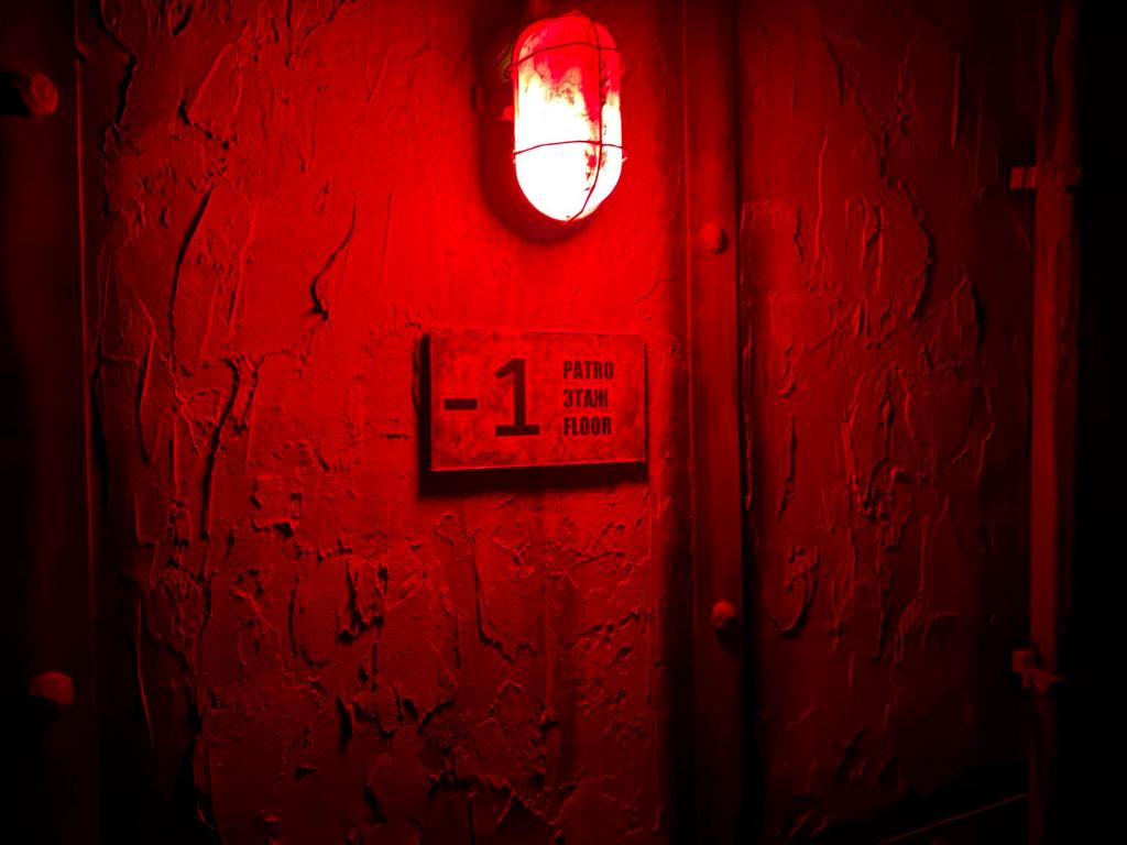 Closeup of an industrial wall lamp. The stuccoed walls have metal strips with rivets. A sign says -1 and the word "floor" in Czech, Russian and English
