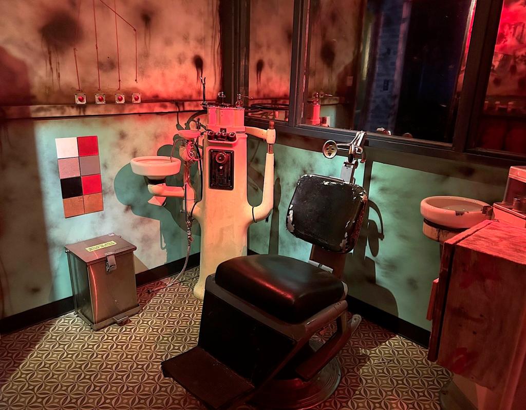A medical exam room with filthy walls. A large black chair with head clamps is in the middle of the room. Other strange equipment fills the corners of the room. The back wall is a big mirror.