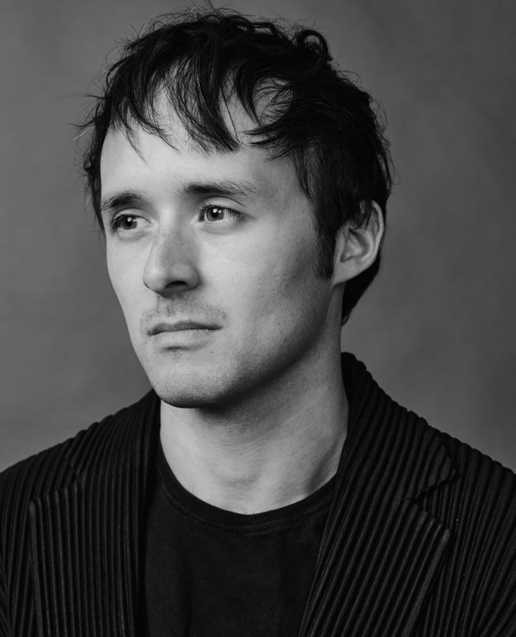 black and white photo of man looking to the left. he has dark hair and is wearing a textured black blazer over a black t-shirt