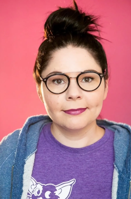 woman wearing glasses, dark hair up in a bun, smiling. She is wearing a blue hoodie over a purple t-shirt