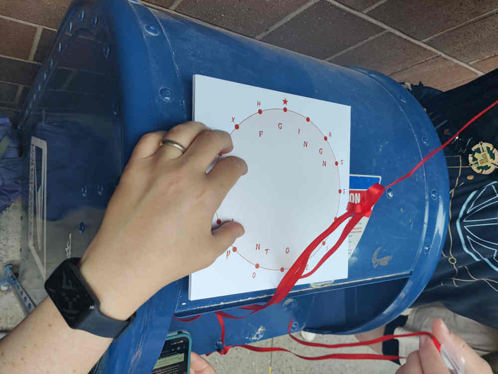 Atop a mail drop box, a hand holds a puzzle being worked on. The puzzle is a white square of posterboard with letters arranged in a circle.