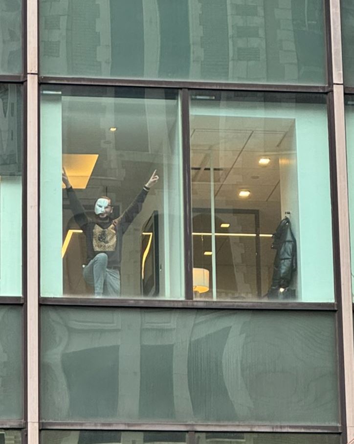man in the window of a high-rise building with a leg propped up, and both hands raised in a V. He is wearing a white mask that covers half of his face diagonally